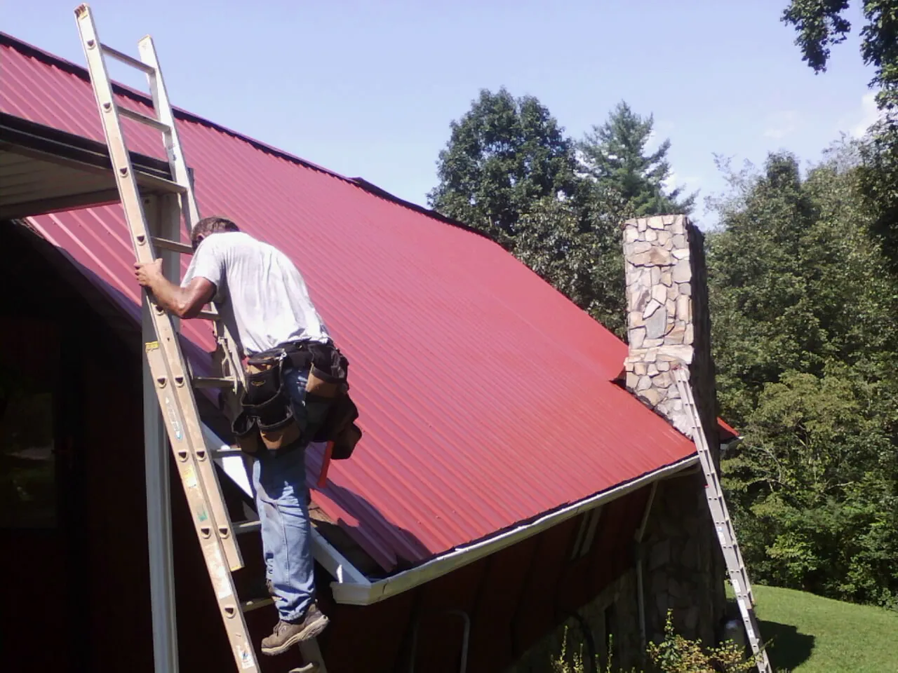 Roofer on a ladder beside a red metal roof and stone chimney — Soffit & Fascia Repair by Roofing Brothers in Kalamazoo